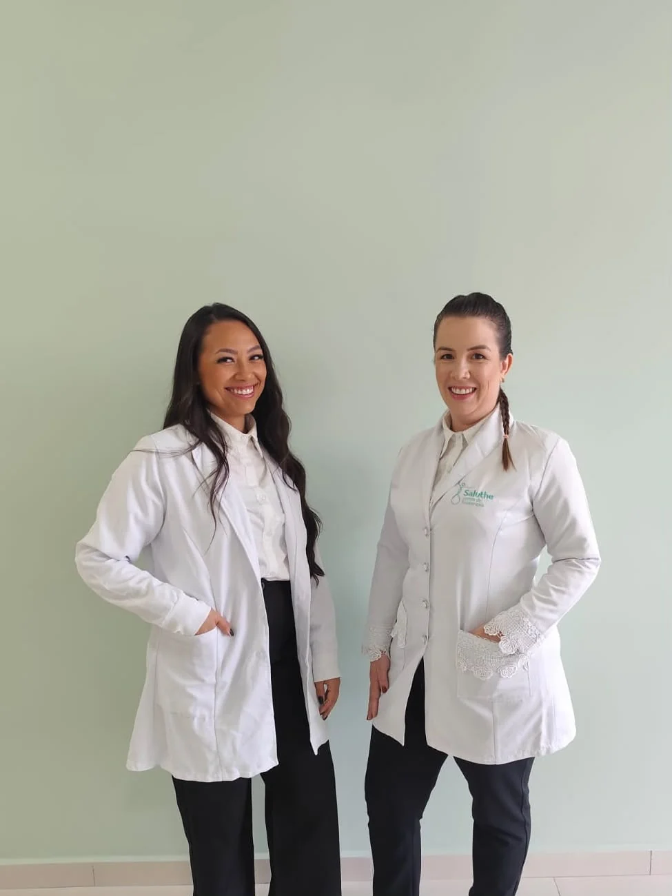 Dra. Rafaela Rigo e Kalyne Tomas, de jaleco, em frente à parede verde-menta da Saluthe