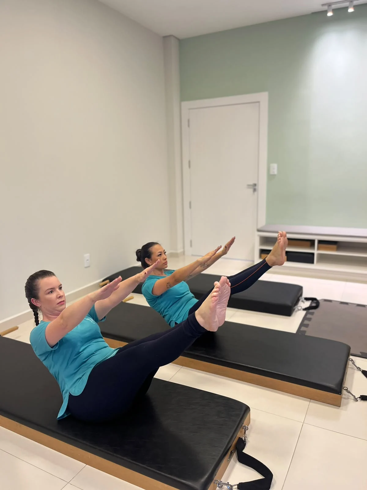 Dra. Rafaela Rigo e uma aluna fazem exercício de Pilates no solo, com as pernas elevadas