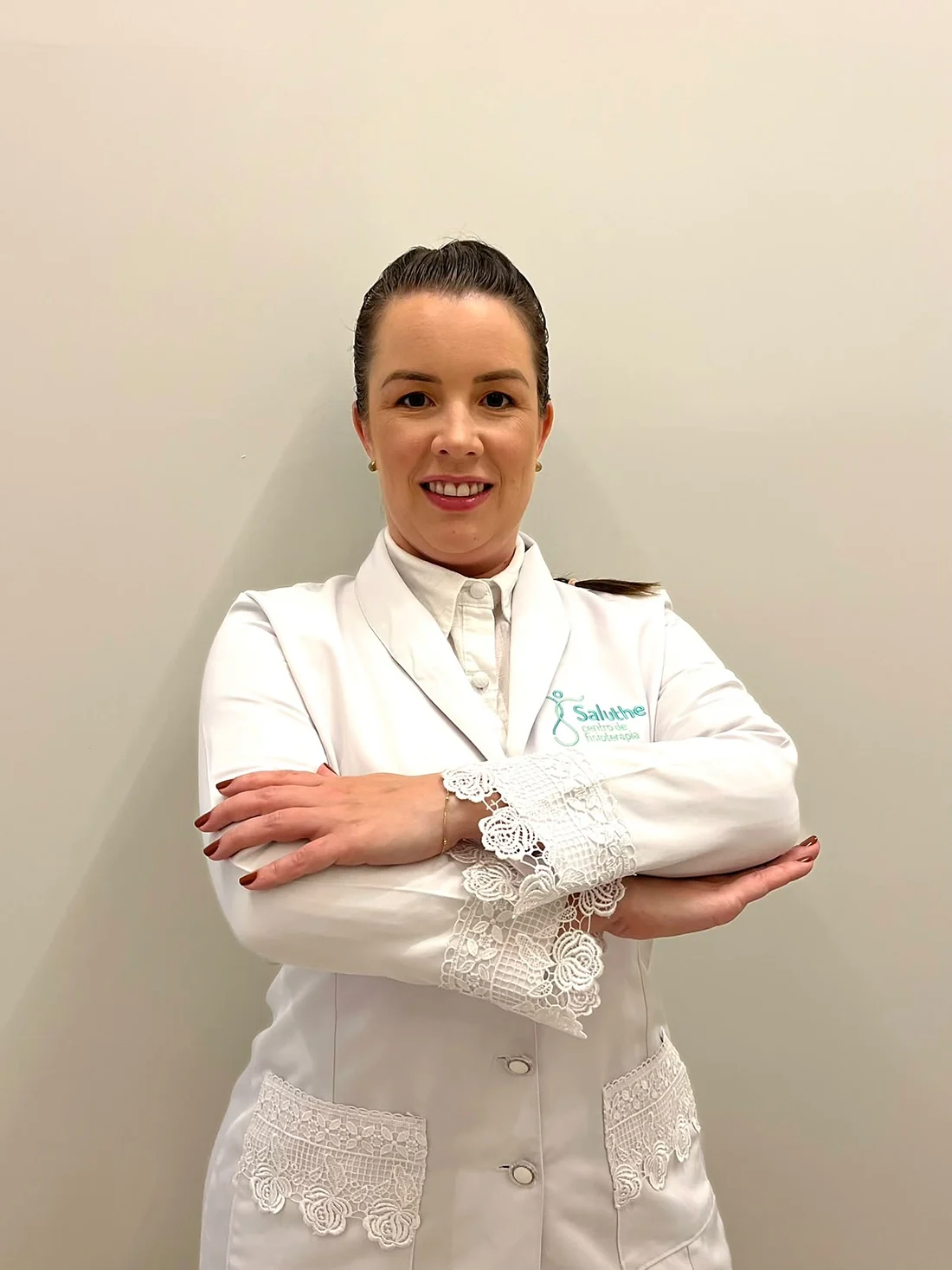 Dra. Rafaela Rigo, fisioterapeuta, de jaleco branco e braços cruzados no consultório da Saluthe